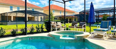Una piscina con cerramiento de cristal, rodeada de sillas de descanso y sombrillas, con vistas a edificios residenciales al fondo.