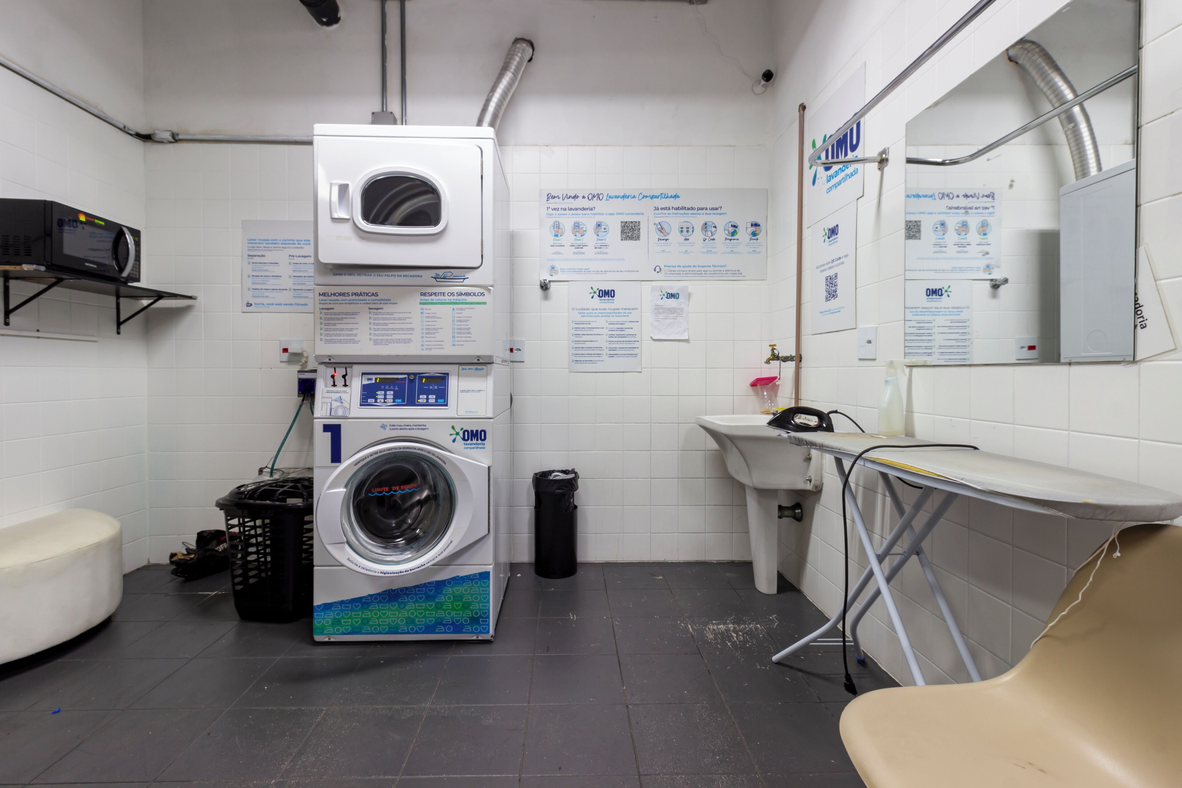 A public restroom with a washing machine, a sink, a mirror, and informational posters on the wall.
