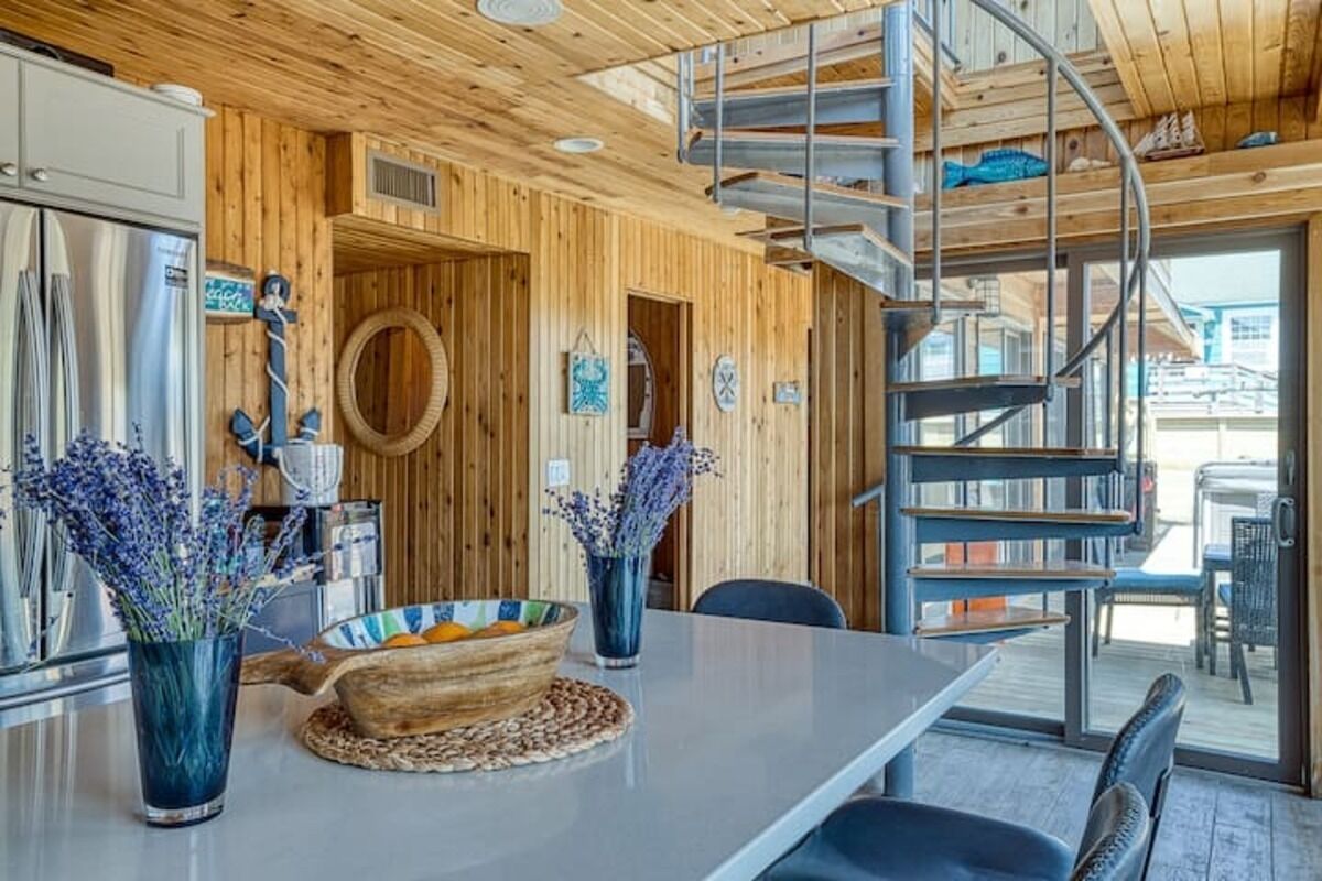 En interiör med träinredning, en spiraltrappa, ett köksområde med ett matbord och utsikt över omgivningen.