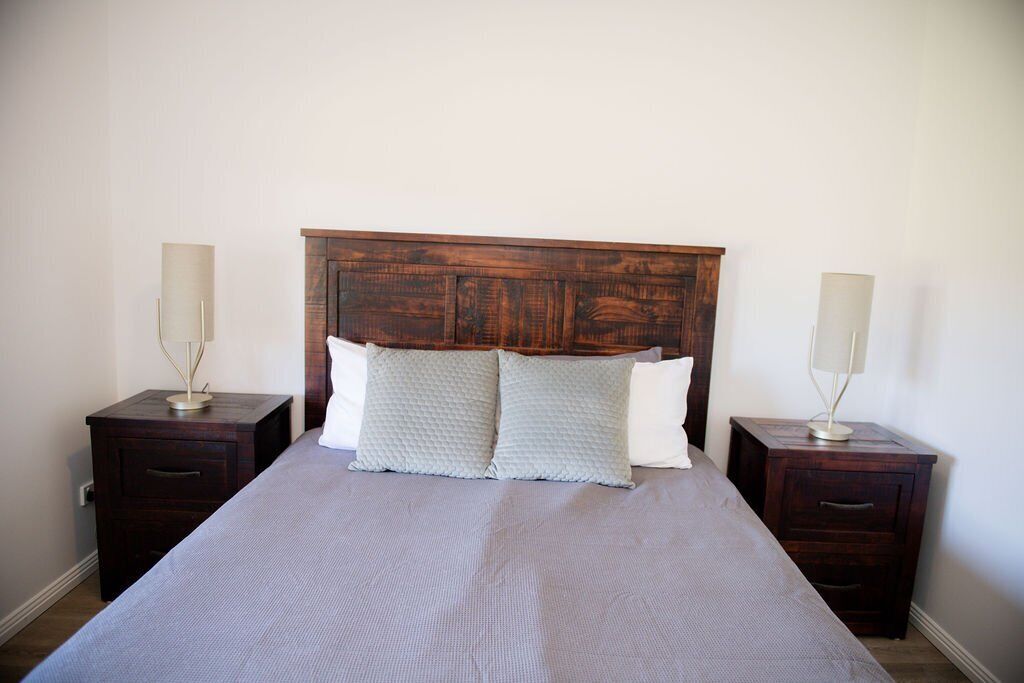 A neatly made bed with a wooden headboard, two bedside tables with lamps, and a plain wall.