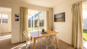 A dining area with a wooden table and clear chairs, a potted plant on the table, and a painting on the wall. - Relax on Rata Cottage 1 (Twizel)