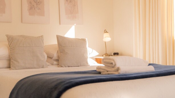A neatly made bed with striped pillows, a blue blanket, and a folded towel on a white mattress. - Relax on Rata Cottage 1 (Twizel)