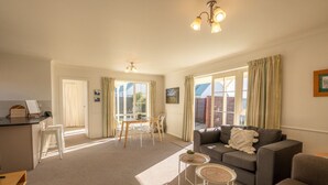 A spacious living room with a grey sofa, a small round table, and a dining area with a wooden table and chairs. - Relax on Rata Cottage 1 (Twizel)
