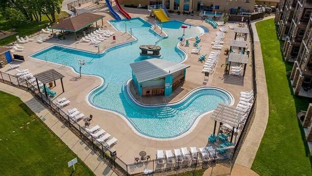 Una piscina con tobogán, tumbonas y un pequeño edificio, rodeada de césped y un edificio al fondo.