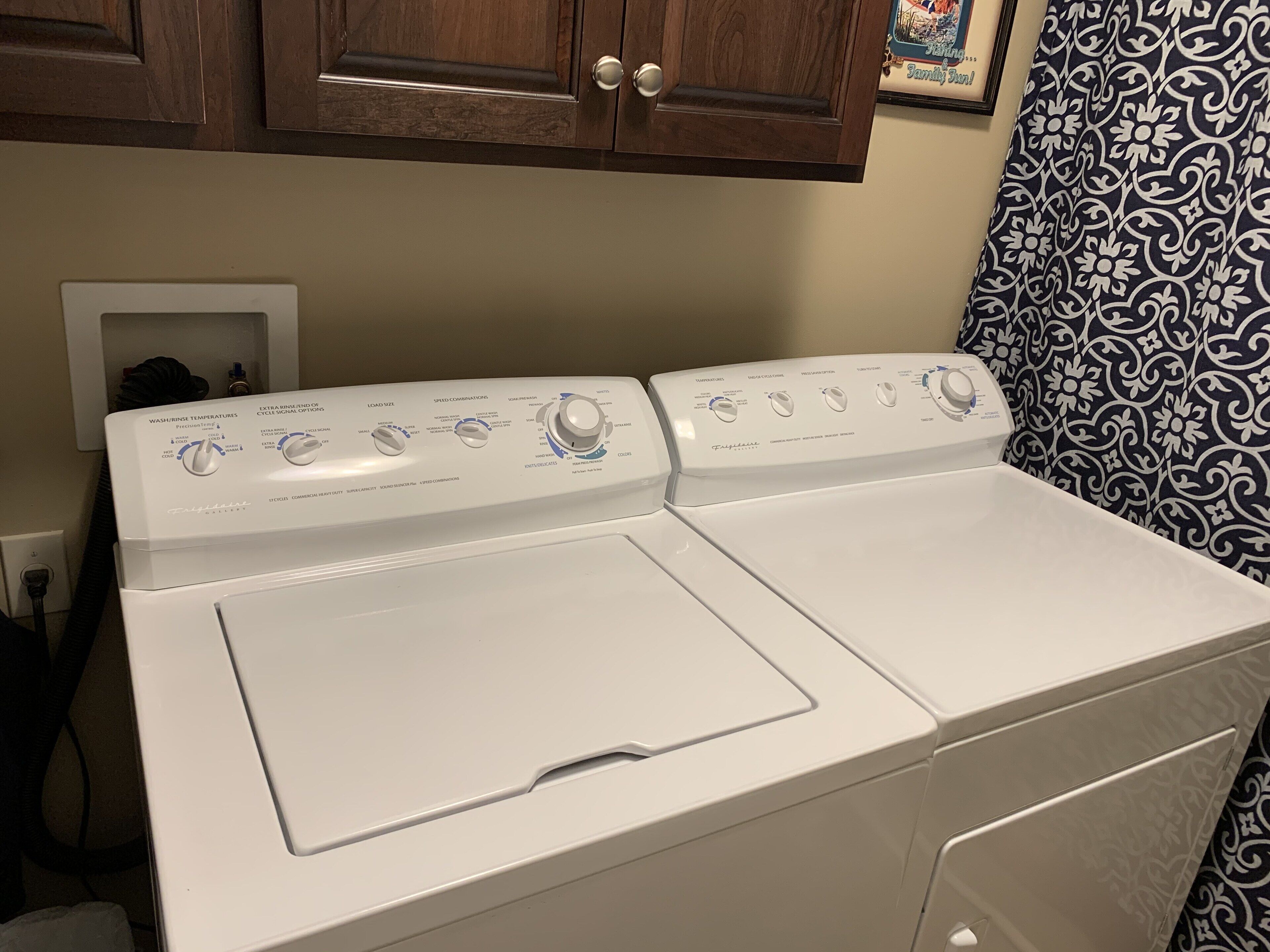 A front-loading washer and dryer in a laundry room with wooden cabinets above and patterned wallpaper on the wall.