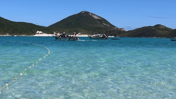 Uma praia com águas turquesa cristalinas, um barco ancorado perto da costa e um cenário montanhoso.