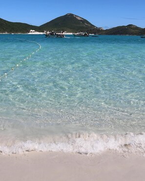 A beach with clear turquoise water, a boat anchored near the shore, and a mountainous backdrop.