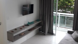 Quarto moderno com cama, TV fixada na parede, prateleira embutida com objetos decorativos e vista para o verde lá fora.