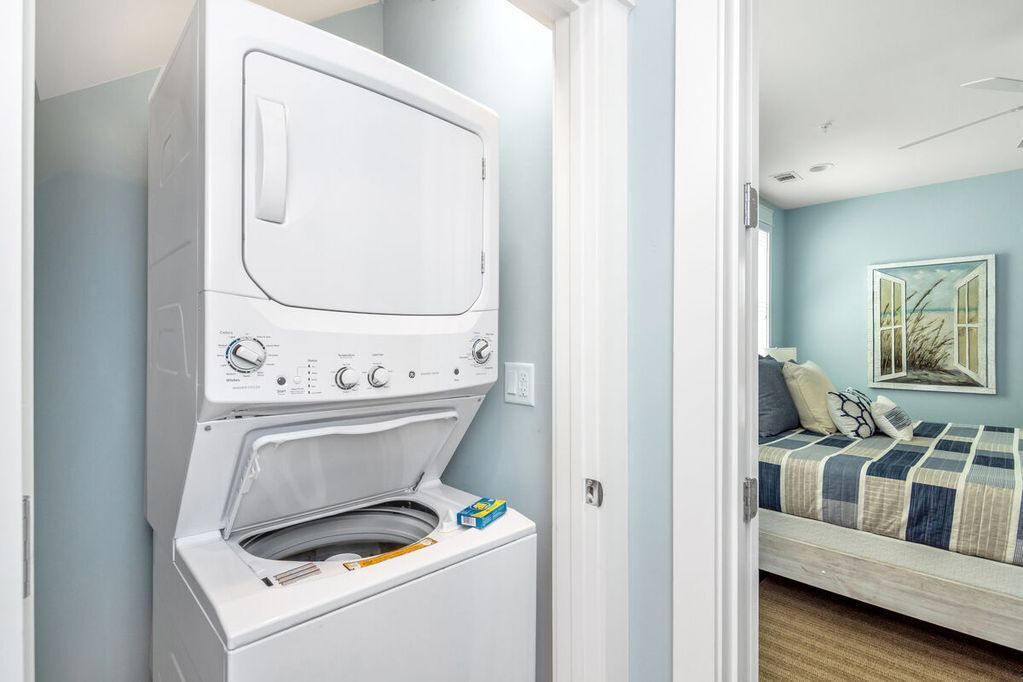 A compact laundry room with a front-loading washer and dryer, a small storage shelf, and a bed with a blue and white checkered bedspread in the adjacent room.