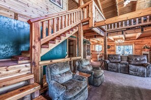 A log cabin interior with a wooden staircase, plush seating, and a chandelier.