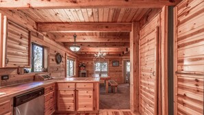 Interior de una cabaña de madera con cocina, comedor y un amplio ventanal.