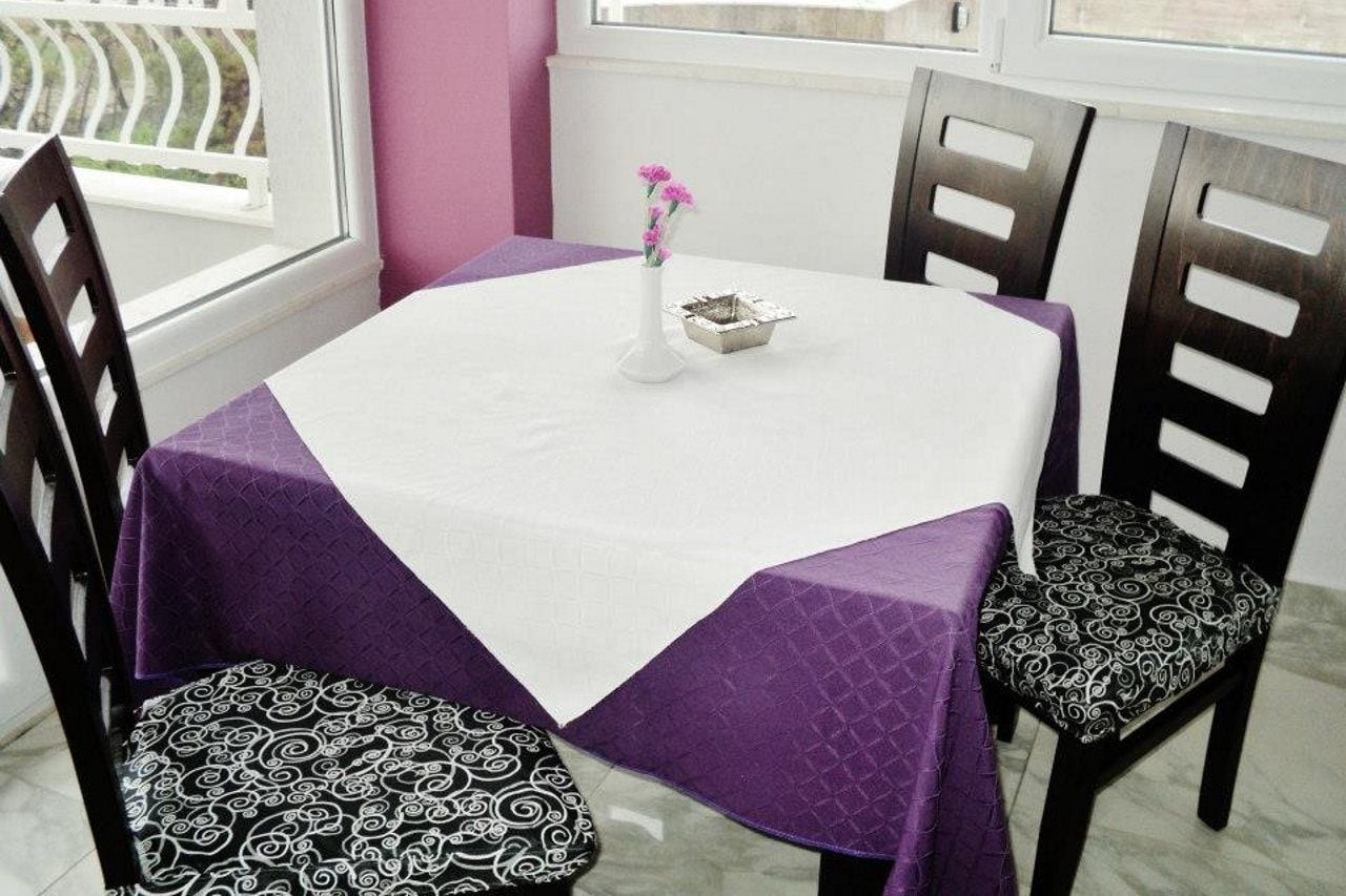 A round table with a white tablecloth and a purple table runner, surrounded by four chairs with patterned cushions.