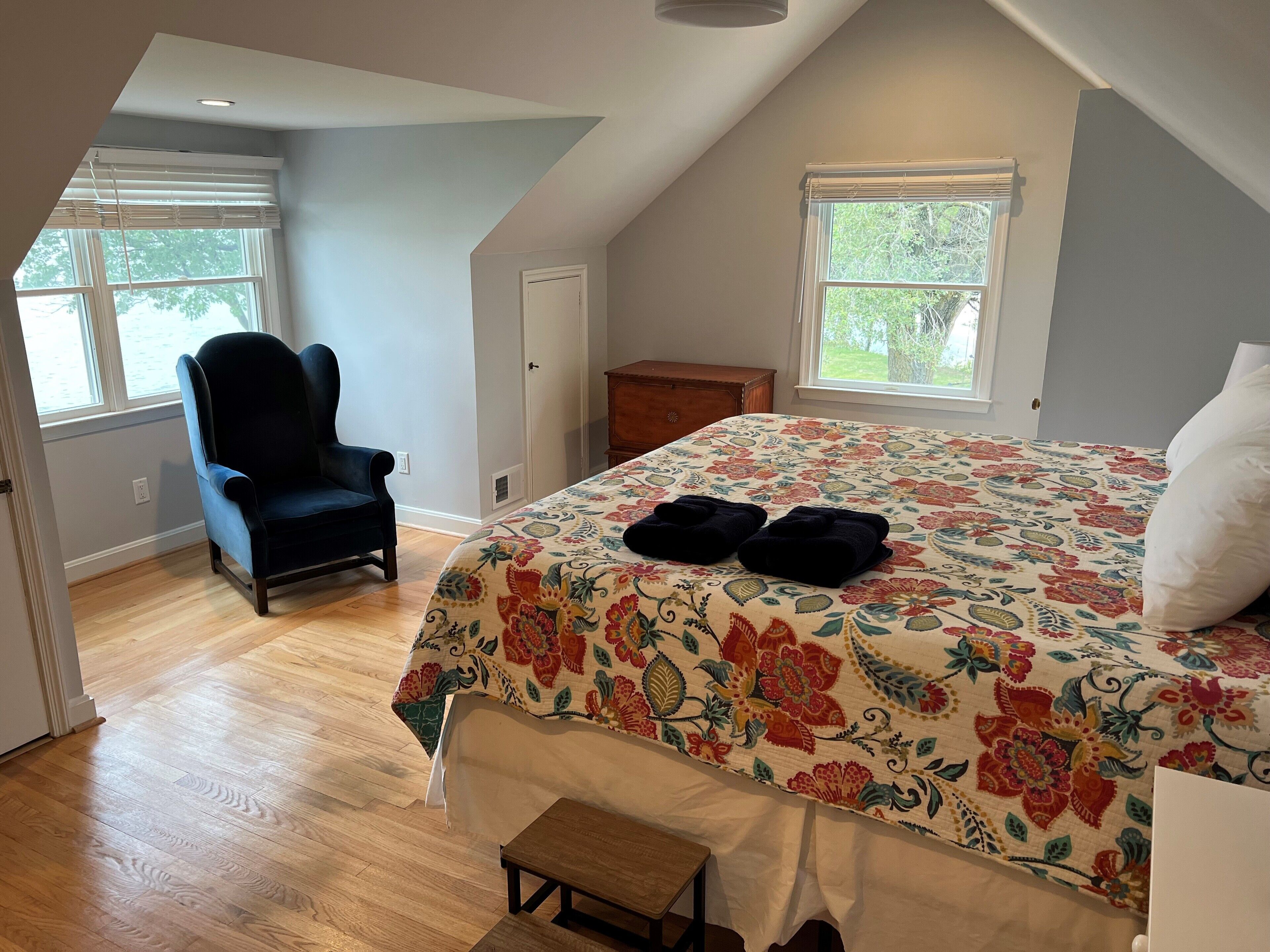 Ein Schlafzimmer mit einem großen Bett, einem Sessel, einer Kommode und zwei Fenstern.
