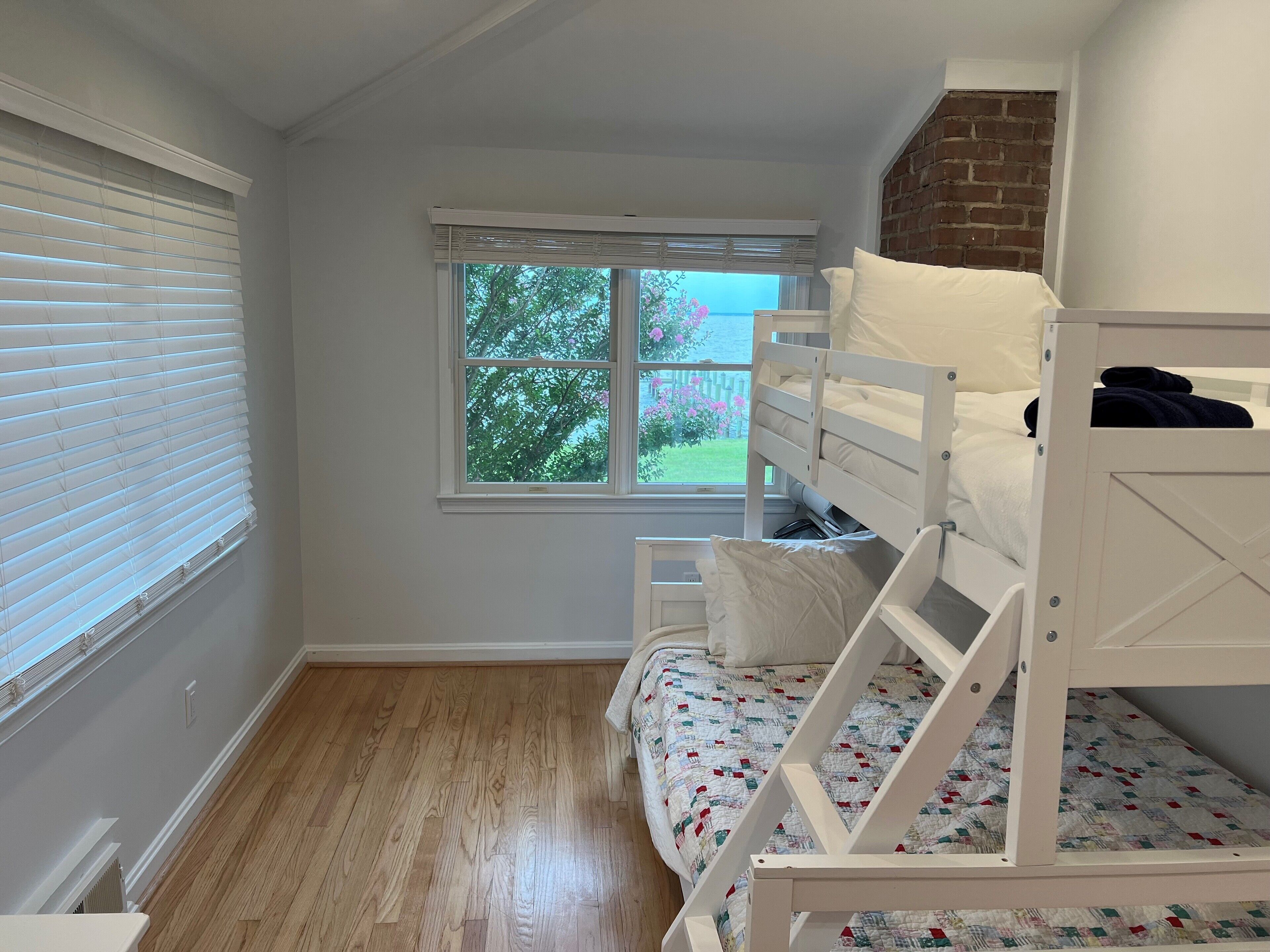 A bedroom with a bunk bed, a window with a view of trees and flowers, and wooden flooring.