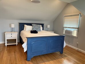 Un dormitorio con una cama azul, mesitas de noche blancas, un cojín estampado y una ventana con persianas.