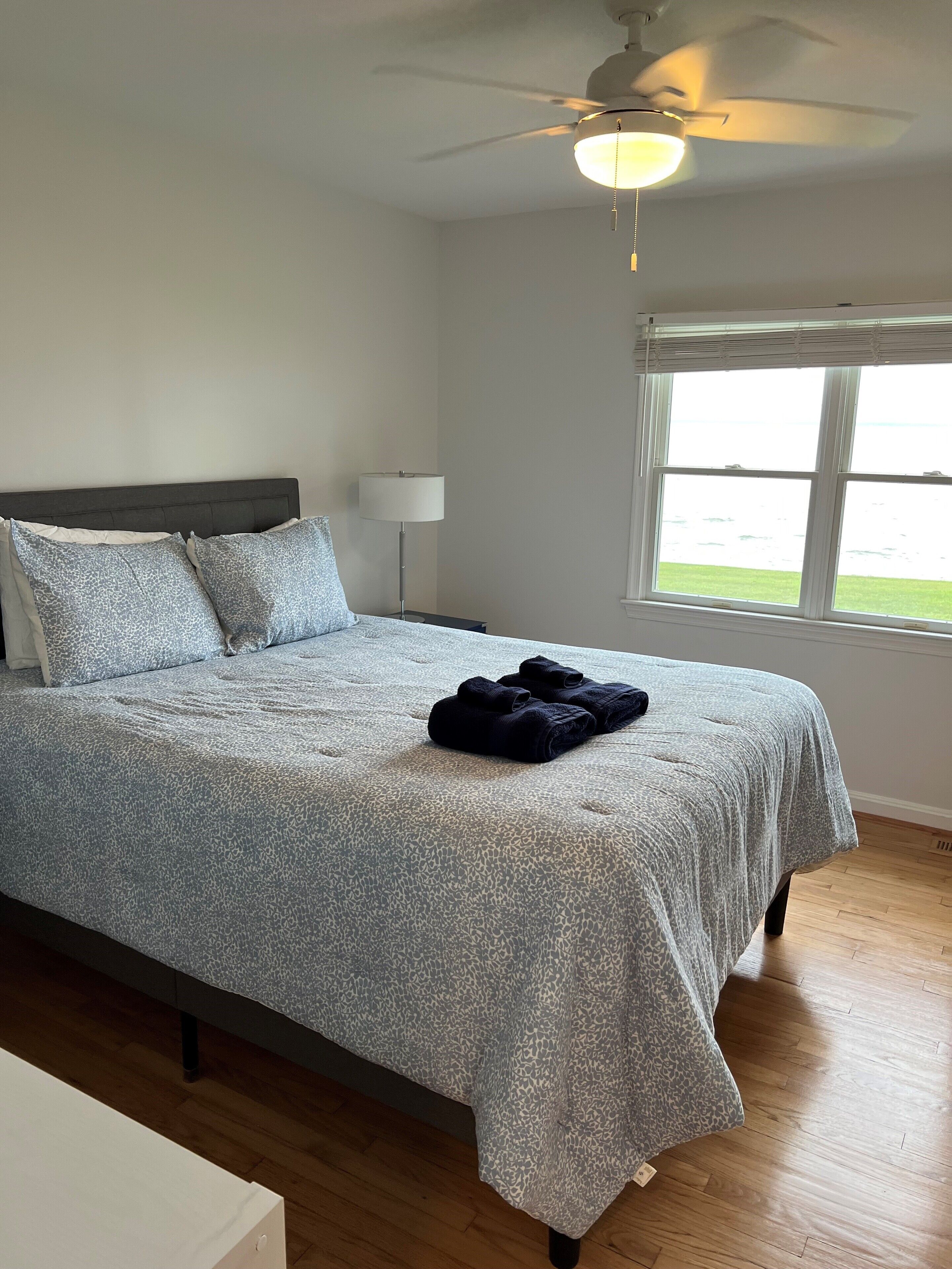 Een slaapkamer met een bed, een plafondventilator, een nachtkastje, een lamp en een raam met uitzicht.