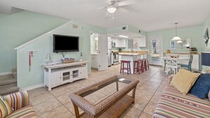 A spacious living room with a television, a sofa, a patterned bench, and a kitchen area in the background.