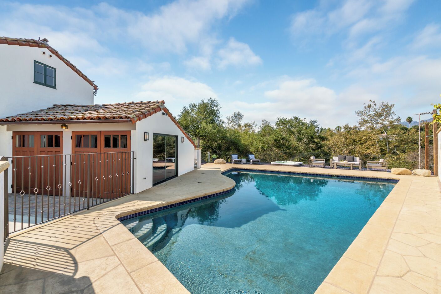 A swimming pool with a tiled deck, a white building with a tiled roof, and a fenced area with outdoor seating.
