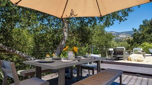 An outdoor dining area with a table set for two, surrounded by wooden chairs and benches, under a large umbrella. - Stunning, Private Home With Pool, Hot Tub, and Mountain Views! (Santa Barbara)
