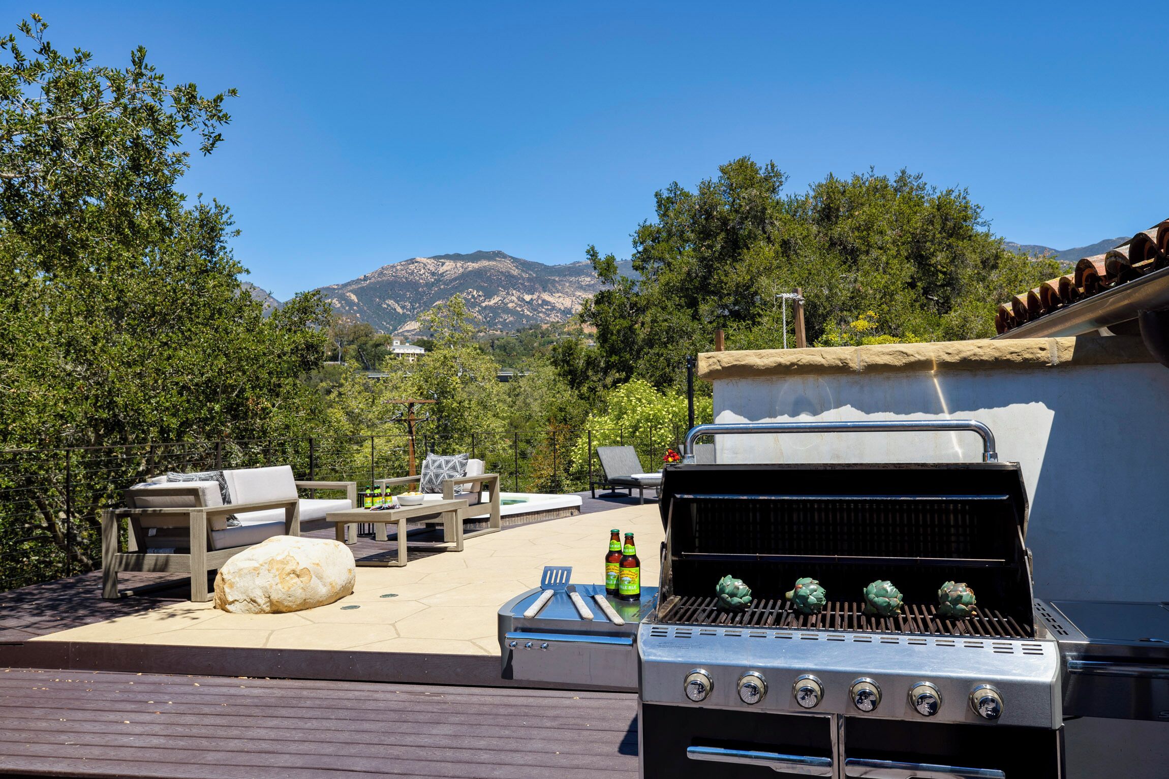 A barbecue grill with two bottles of beer, a large rock, and outdoor furniture on a patio with a mountain view.