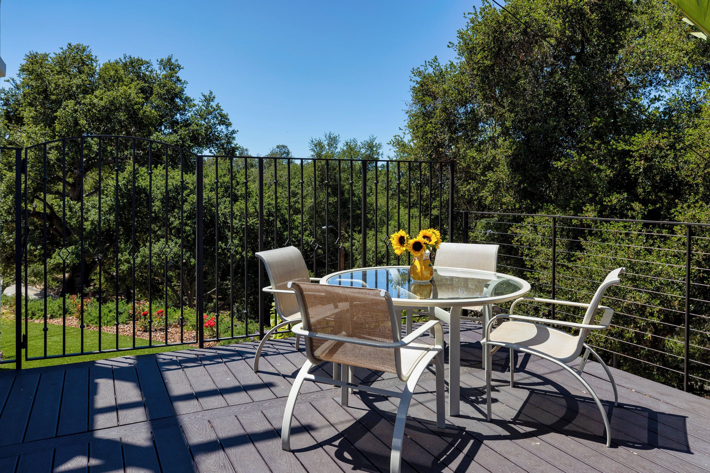 A deck with a round glass table and four chairs, surrounded by a black metal fence.
