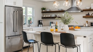 Une cuisine moderne avec un réfrigérateur en acier inoxydable, des chaises noires, un îlot central agrémenté d’une plante et une bouteille de vin.