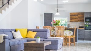 A modern living room with a grey sofa, wooden dining table, and a kitchen area with dark cabinets and a built-in oven. - Villa Cedar with large swimming pool and summer kitchen (Gostinjac (Krk))