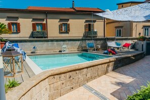 A swimming pool with lounge chairs and a pergola, surrounded by a stone wall and a building with balconies.
