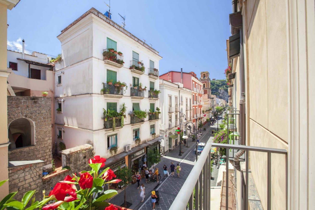 A narrow street with buildings on both sides, balconies with plants, and pedestrians.