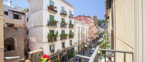 A narrow street with buildings on both sides, balconies with plants, and pedestrians.