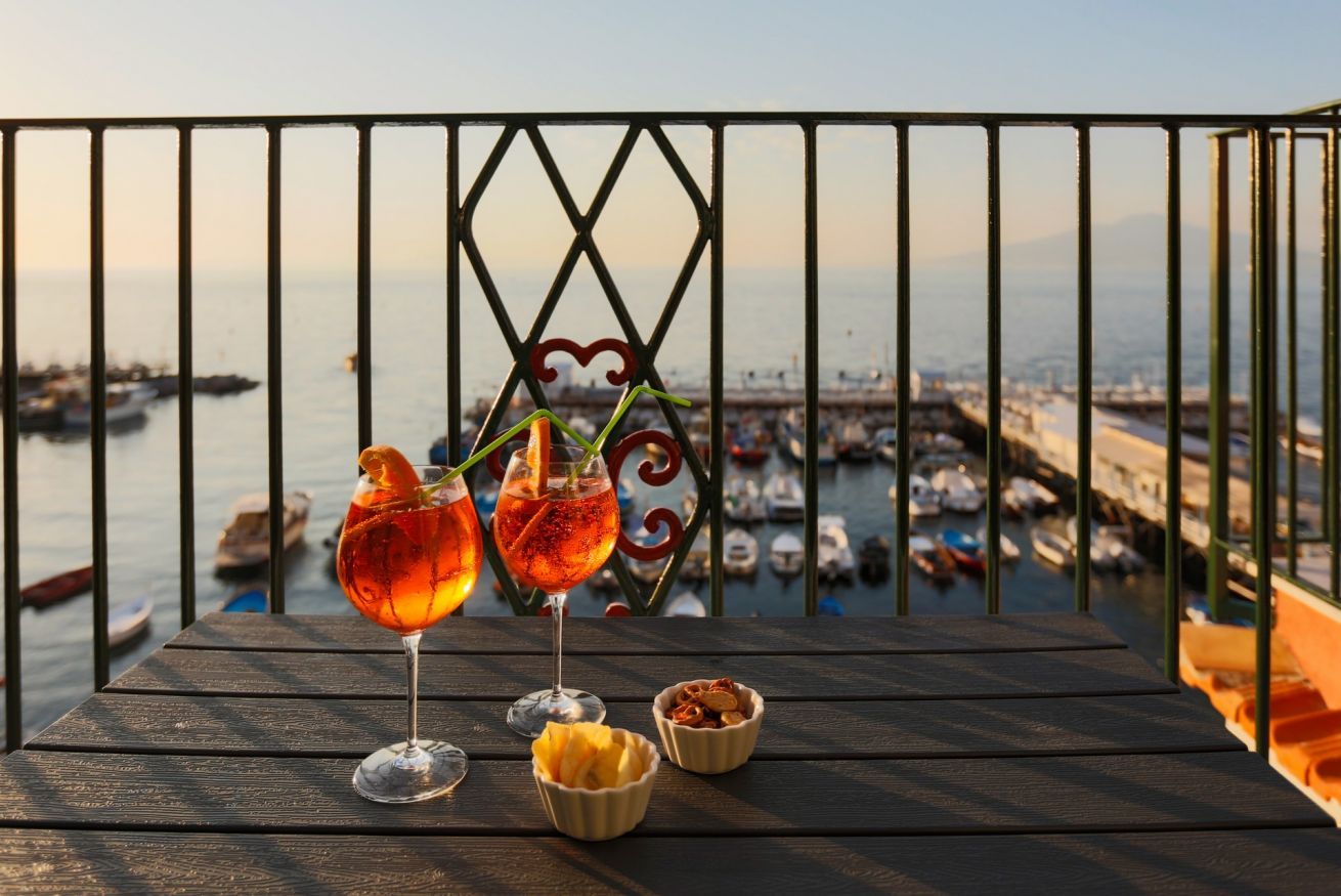 A balcony with two cocktails and snacks, overlooking a marina with boats.