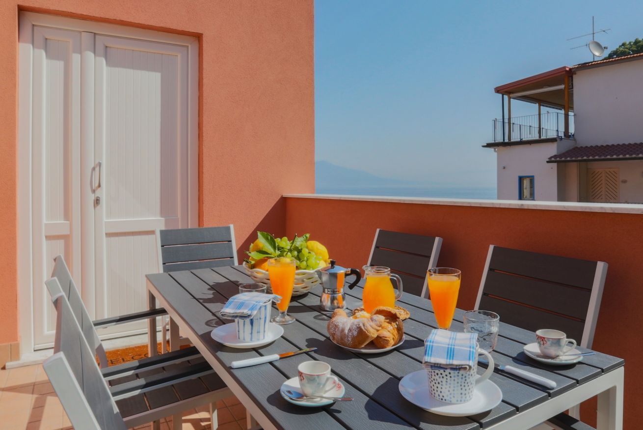 Une table dressée pour le petit-déjeuner, avec du jus d’orange, des viennoiseries et un panier de fruits, sur un balcon offrant une vue imprenable.