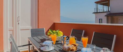 A table set for breakfast with orange juice, pastries, and a fruit basket on a balcony with a view.