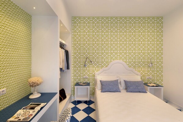 A bedroom with a patterned green and white wall, a white bed with patterned pillows, a blue and white checkered floor, and a small bedside table with a lamp.