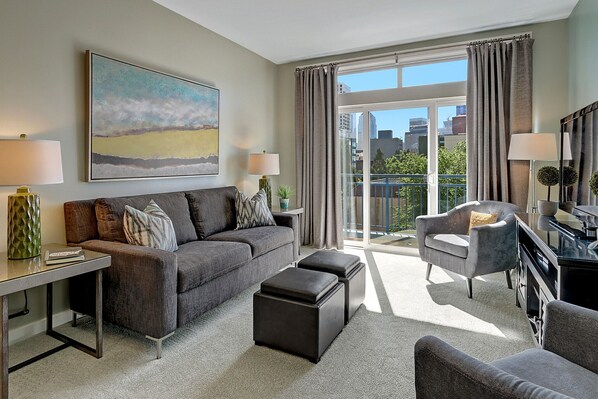 A modern living room with a grey sofa, a coffee table, and a side table with a lamp. There is a large window with curtains and a view of the city. - City ★ Water View Oasis with Pool & Gym near Cruise in Heart of Seattle (Seattle)