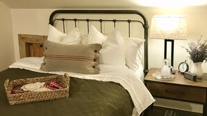 Una cama bien tendida con una colcha verde, almohadas blancas y una cesta trenzada con un paño de cuadros rojos y blancos. Hay una mesita de noche con una lámpara, un reloj y un jarrón con flores.