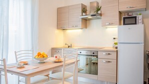 A modern kitchen with light wood cabinets, a stainless steel oven, a microwave, and a refrigerator. There is a dining table with chairs and a bowl of fruit.