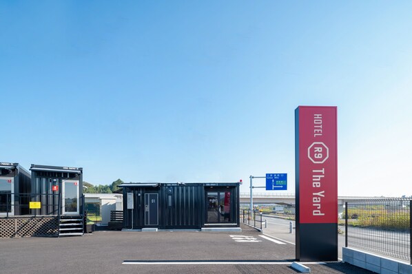 A modern hotel entrance with a red sign, a small building with a glass door, and a clear sky. - HOTEL R9 The Yard Ami (Ami)