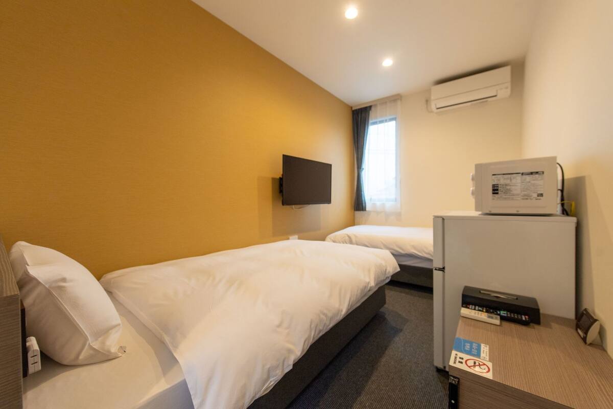 Hotellihuone, jossa on kaksi erillistä sänkyä, seinälle asennettu taulutelevisio, ilmastointilaite ja jääkaappi.