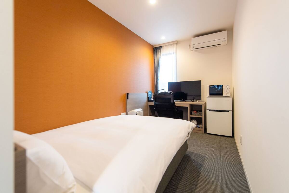 Hotellihuone, jossa on sänky, työpöytä tietokoneella, jääkaappi ja ilmastointilaite.