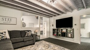 Una sala de estar moderna con un sofá seccional gris, un tapete abstracto grande, un televisor de pantalla plana montado en la pared y una estantería con objetos decorativos.