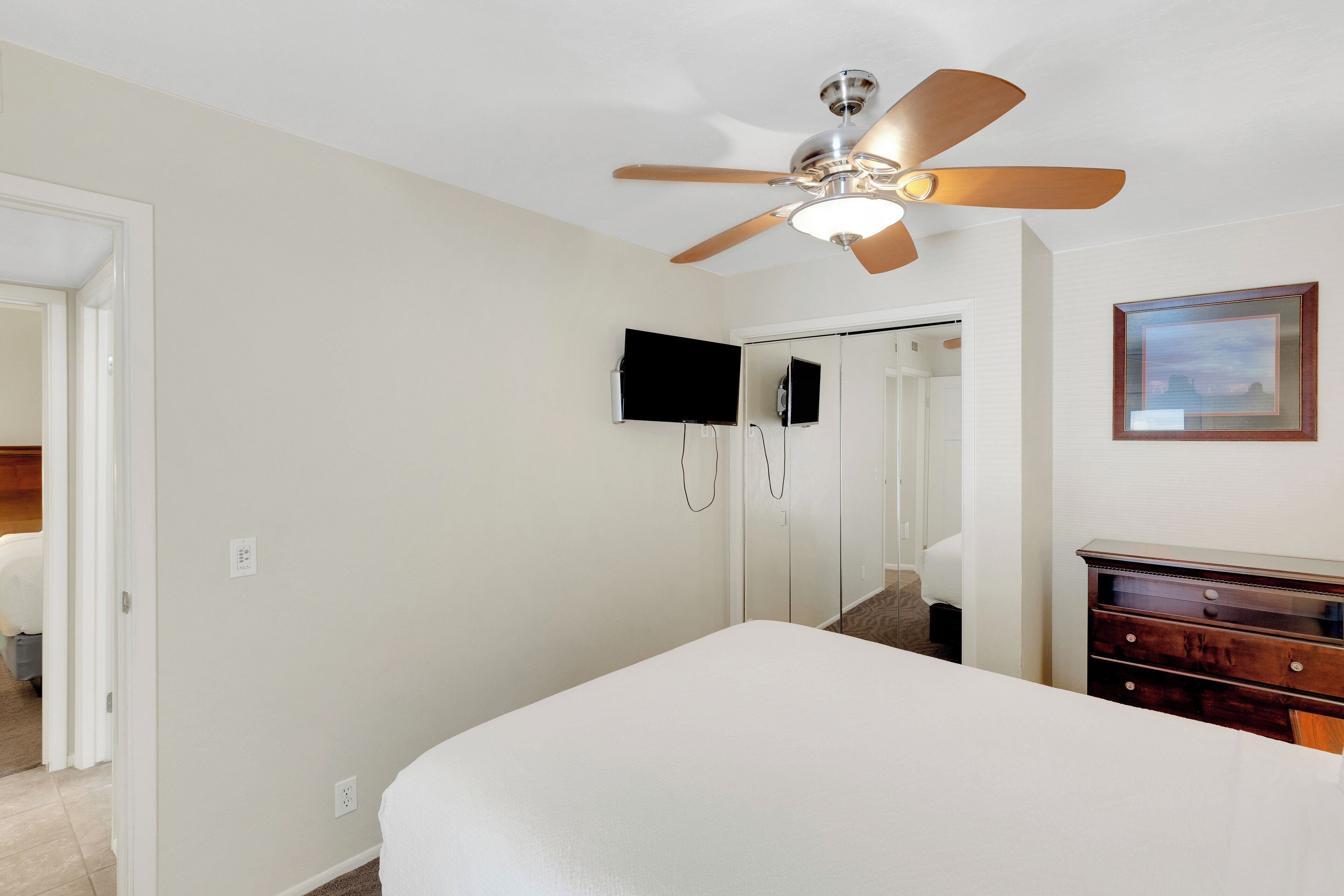 A bedroom with a bed, a ceiling fan, a dresser, a TV, and a door leading to another room.