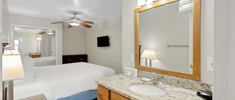 A hotel room with a bed, a large mirror, a vanity with a sink, and a ceiling fan.