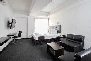 A modern hotel room with a bed, a desk, a chair, a sofa, and a TV mounted on the wall.