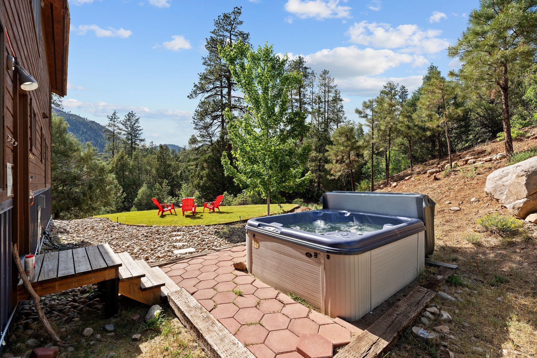 Een hottub in een achtertuin met een houten terras, omringd door bomen en uitzicht op een berg.