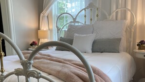 A bedroom with a white bed, a white headboard, and a window with white curtains.