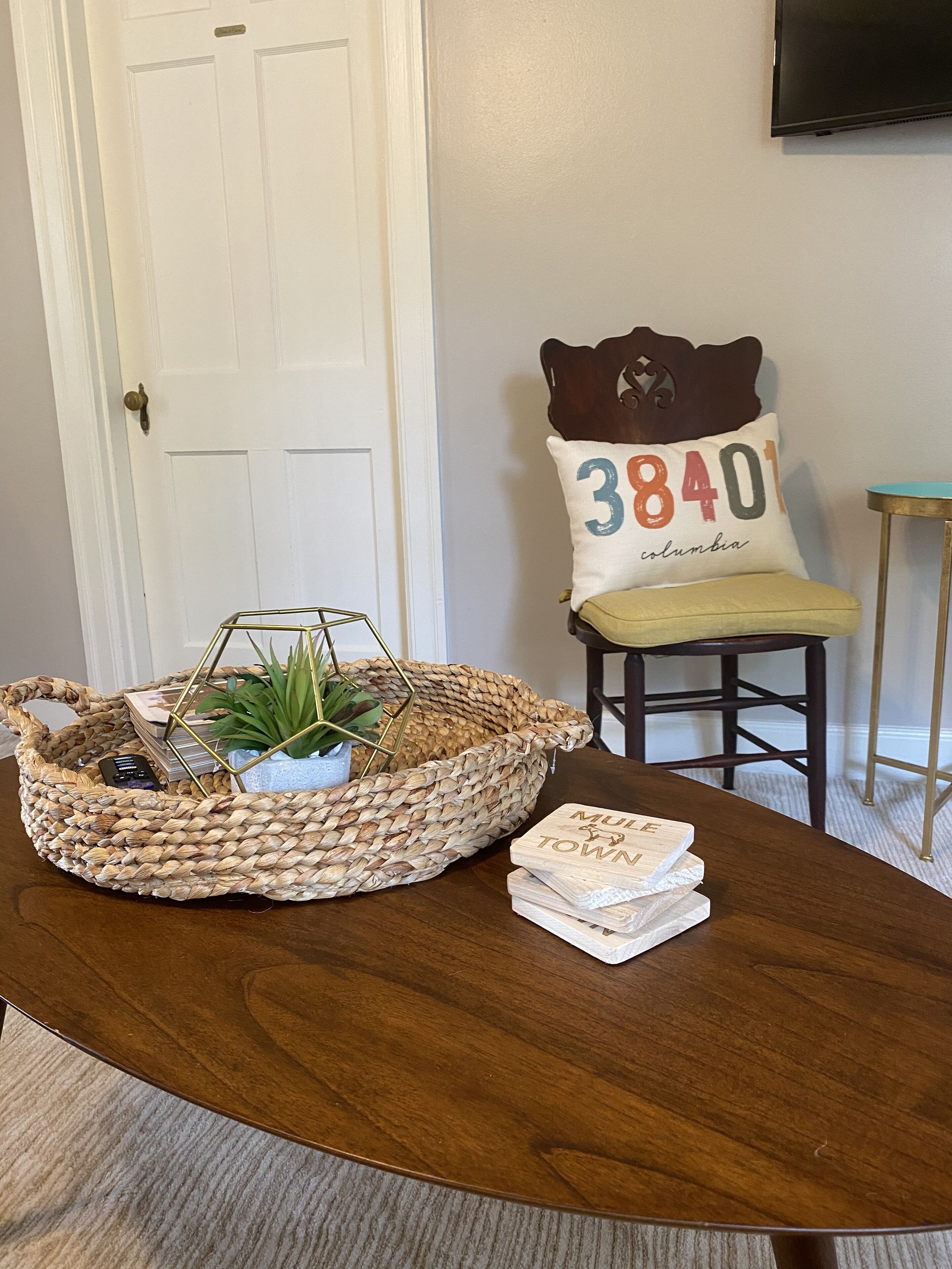 Een houten tafel met een gevlochten mand met een plant en boeken, een stoel met een kussen met de tekst '38401 Columbia', en een bijzettafeltje met een televisie aan de muur.