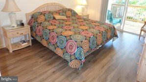 A bedroom with a wooden bed, two bedside tables with lamps, a framed picture on the wall, and a view of the outdoors through a sliding glass door. - Gorgeous 3 Bedroom Townhouse, Bethany Beach, steps from pool and fitness center (Bethany Beach)