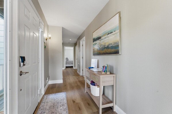 A hallway with a wooden floor, a white door, a desk with a basket of pens, and a painting on the wall.