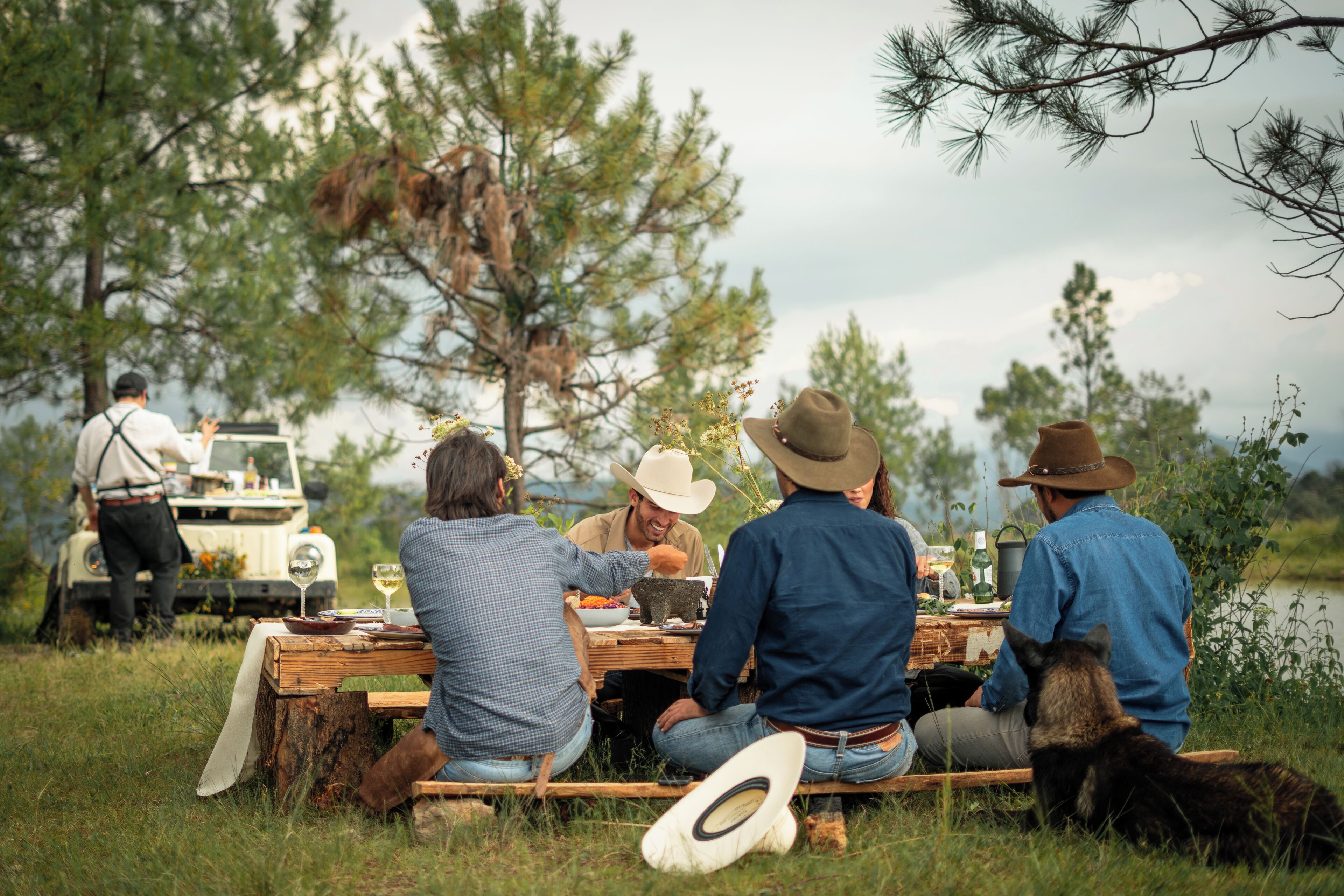 Un grupo de personas cenando al aire libre, con una mesa dispuesta para una comida, rodeadas de naturaleza.